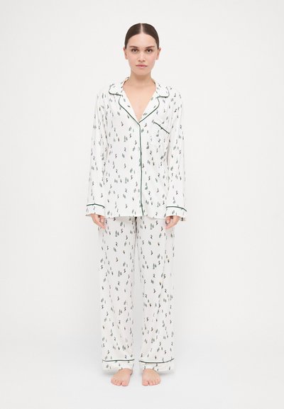 Set di pigiama bianco con motivo a pini sparsi, rifiniture verdi, e una camicia con bottoni e tasca, abbinata a pantaloni larghi.