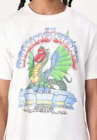 White t-shirt featuring a colorful graphic of a dragon with green wings and a red tongue, above "The Rolling Stones" text and a venue illustration.