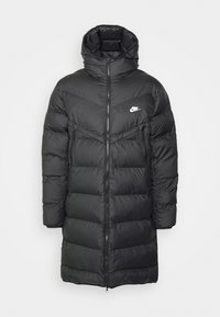 Svart puffjacka med hög krage och dragkedjestängning. Har en Chevron-design, två sidofickor och en liten vit logotyp på bröstet.