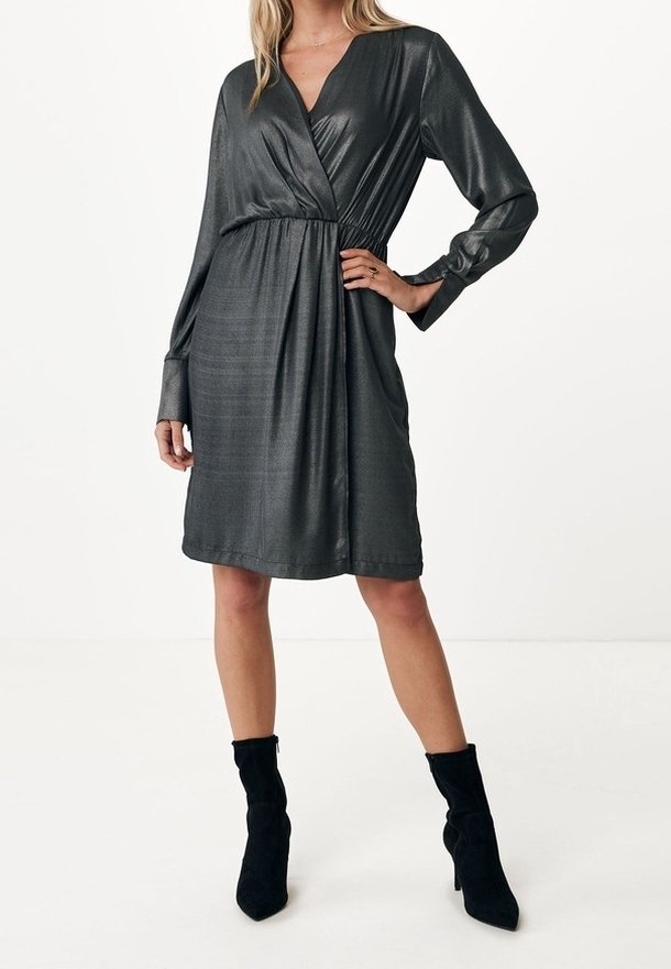 Femme portant une robe portefeuille métallique charbon à manches longues et longueur genou, avec des bottines en daim noir, debout, une main sur la hanche.