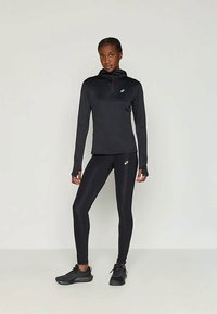 Naine seisab mustas spordihigis, retuusides ja mustades jooksujalatsites, ühtlase vaale tausta ees.