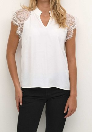 Vrouw draagt een witte blouse met kanten kapmouwen en zwarte broek, staand tegen een effen lichte achtergrond.