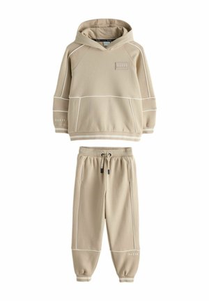 Bež jopič s kapuco in ujemajoče se trenirke iz mehkega blaga, z belimi robovi in "BAKER" blagovno znamko na obeh kosih.