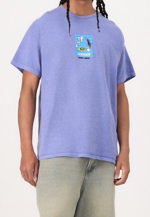 Osoba ubrana w lawendowy T-shirt z niebieską kratą przedstawiającą trzy karpie koi oraz napis „sensei LEARN + GROW” na piersi.