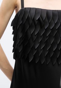 Robe noire bustier avec des détails de tissu superposés en forme de pétales couvrant le haut du corsage, laissant les épaules et les bras nus.