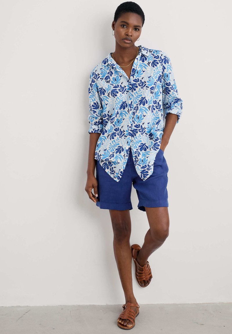 Camicia a fiori con bottoni in azzurro chiaro e blu navy, con maniche lunghe arrotolate. Abbinata a pantaloni corti blu navy e sandali marroni con cinturini.