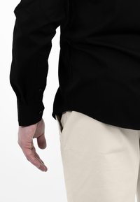 Zwarte langwerpige overhemd met knopenmanchetten, met een gladde textuur en subtiele glans, gecombineerd met lichtbeige pantalons.