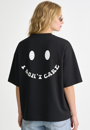 Blonde woman wearing black oversized t-shirt with white smiley face and "I DON'T CARE" text on the back, paired with light blue jeans.