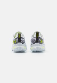 Baskets de sport blanches avec un dessus texturé noir et gris, présentant une accentuation jaune vif et une languette au talon.
