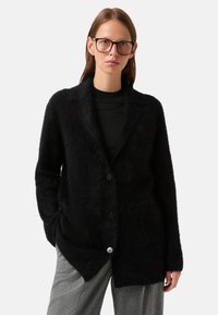 Černý chlupatý cardigan s klopovým límcem, dlouhými rukávy a zapínáním na dva knoflíky. Nosený přes černou košili v kombinaci se šedými kalhotami.