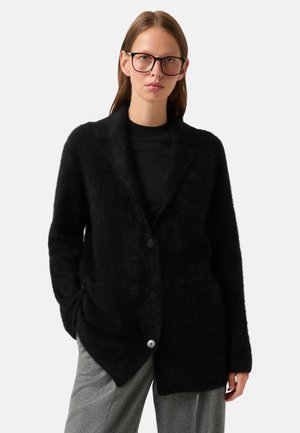 Zwarte donzige cardigan met een gelapte kraag, lange mouwen en sluiting met twee knopen. Draag over een zwart shirt, gecombineerd met een grijze broek.