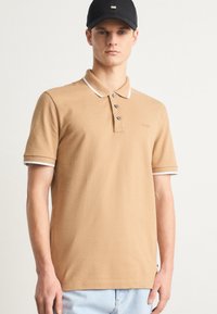 Tan polo majica s kratkimi rokavi, kontrastnim belim ovratnikom in poudarki na rokavih, tri gumbi in diskretno blagovno znamko na prsih.