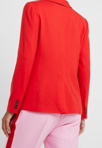 Punainen bleiseri, jossa on jämäkkä kaulus, istuva leikkaus ja napitetut hihansuut. Kangas näyttää teksturoidulta ja se on esitelty vaaleanpunaisien housujen kanssa.