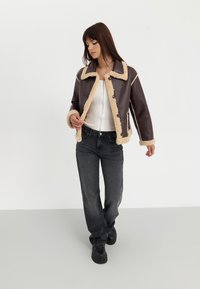 Brun kunstlær jakke med beige faux pels trim, knappelukking og kragehals, kombinert med mørke jeans og svarte sko.