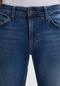 Jeans dżinsowe w średnim niebieskim kolorze z klasycznym krojem. Do cech należą zapięcie na guzik, kieszenie z przodu oraz delikatne detale przeszycia.