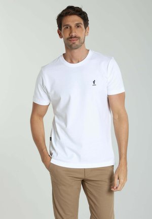 Homme portant un t-shirt blanc à manches courtes avec un petit logo sur la poitrine, pantalon beige, debout avec une main dans la poche sur fond uni.