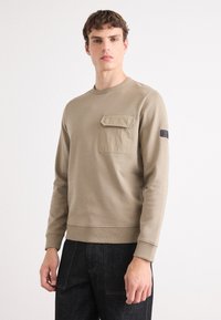 Sweatshirt bege em tecido macio, com um bolso frontal e punhos canelados. Combinado com jeans em denim escuro. Design simples de gola redonda.