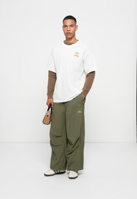 Hvid t-shirt med korte ærmer over en brun langærmet skjorte, parret med løse olivengrønne bukser. Tilbehør omfatter en lille taske. Hvide sneakers med striber.