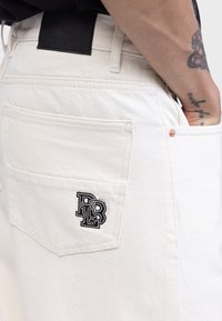 Pantalon en denim blanc avec un logo noir brodé sur la poche arrière, doté d'un tissu texturé et de rivets en cuivre.