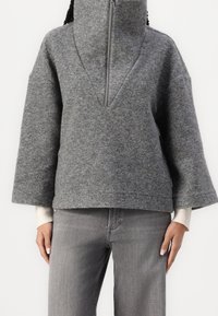 Sudadera de forro polar gris con cuello alto y media cremallera, que presenta mangas anchas y un ajuste holgado. Combinada con jeans grises.