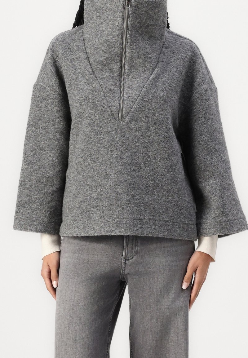 Sudadera de forro polar gris con cuello alto y media cremallera, que presenta mangas anchas y un ajuste holgado. Combinada con jeans grises.