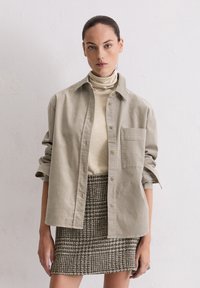 Light gray button-up shirt with a chest pocket, layered over a beige turtleneck. Paired with a woven checkered skirt in brown tones.
