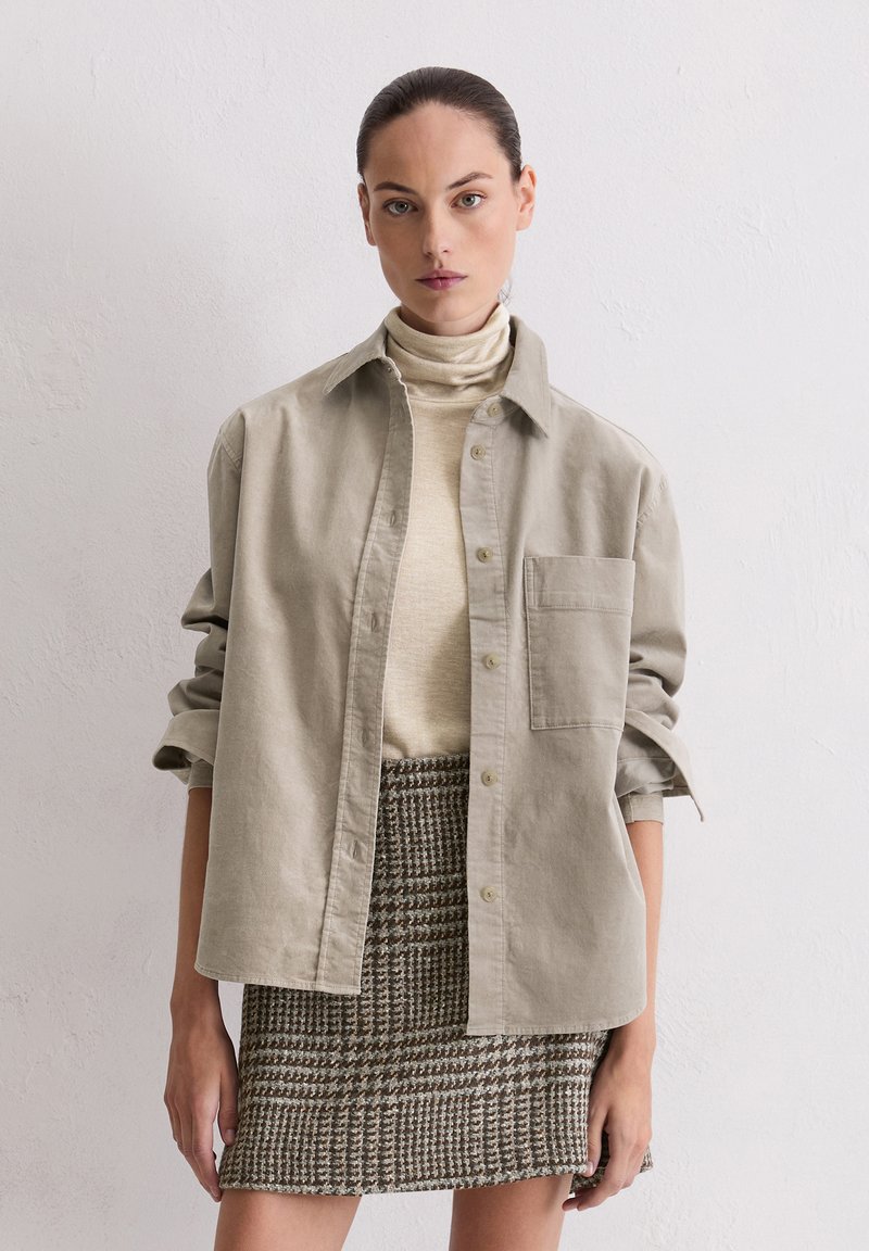 Light gray button-up shirt with a chest pocket, layered over a beige turtleneck. Paired with a woven checkered skirt in brown tones.