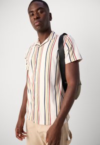 Chemise à manches courtes et à boutons avec une base blanche, présentant des rayures verticales rouges, dorées et bleu marine. Tissu lisse et design classique de col.