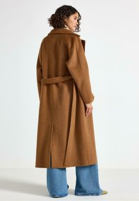 Cappotto in misto lana marrone con vita regolabile, maniche lunghe e apertura sul retro, abbinato a jeans blu svasati e calzature verdi.