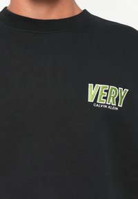 Μαύρο πουλόβερ με στρογγυλή λαιμόκοψη, με πράσινο και λευκό κεντητό κείμενο "VERY" και μικρότερο "Calvin Klein" από κάτω στο στήθος.