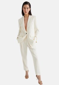 Abito su misura color avorio con scollatura a V profonda, ampi revers, due tasche frontali e pantaloni dritti, abbinato a sandali con tacco alto e fasce.