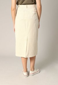 Jupe en velours côtelé de couleur crème, longueur genou, avec une fente à l'arrière, dotée de deux poches arrière et d'une taille ajustée. Associée à des baskets blanches.