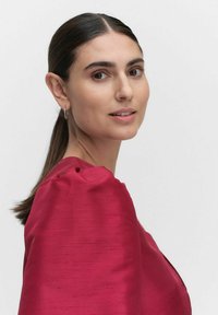 Rød satin top med puffede ærmer, der har en struktureret overflade. Modellen har glat hår og minimalistiske accessories, sat mod en hvid baggrund.