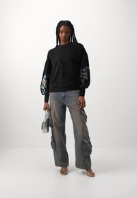 Svart sweatshirt med ballongermar som har blomsterbroderi; kombinert med falmet cargo-jeans og transparente hæl-sandaler. Holder en sølvveske.