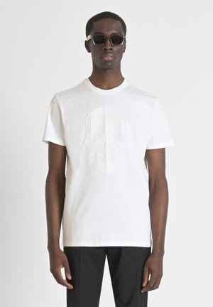 Witte katoenen t-shirt met een verhoogd doodshoofdontwerp in het midden. Model draagt een zonnebril, met aansluitende zwarte broek die de look compleet maakt.