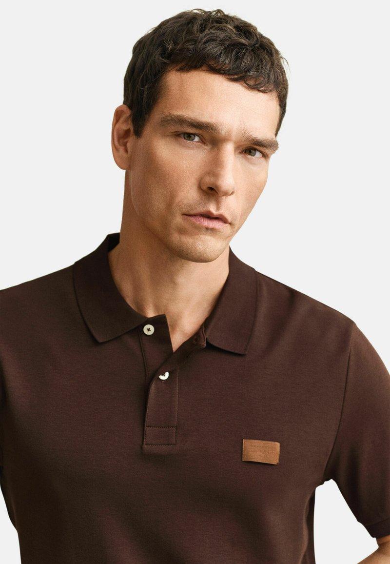 Jonge man met kort bruin haar, gekleed in een donkerbruin poloshirt met een kleine beige patch op de borst, kijkt recht vooruit met een neutrale uitdrukking.