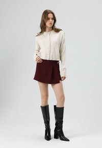 Beige cropped cardigan with buttons, maroon skirt with pleat detail, and knee-high black shiny heeled boots. Simple design, smooth texture.