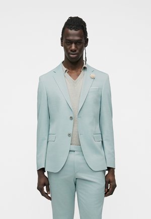 Mann mit dunkler Haut und geflochtenem Haar trägt auf weißem Hintergrund einen hellblauen Anzug und einen beigen V-Ausschnitt-Pullover.