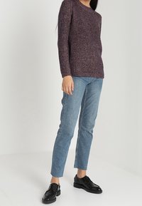 Strickpullover in Dunkellila mit strukturiertem Muster, kombiniert mit hellblauen Jeans mit geradem Bein und schwarzen Schnürschuhen, stehend vor weißem Hintergrund.