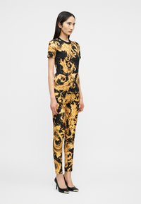 Γυναίκα όρθια, φορόντας μαύρο και χρυσό μπλουζάκι με κοντά μανίκια και παντελόνι σε μπαρόκ μοτίβο, με μαύρες ψηλοτάκουνες μυτερές γόβες.