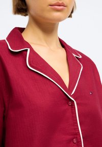 Burgundská pyžamová blúzka s bielym lemovaním, s výstrihom do V, zapínaním na gombíky a vyšitým logom na ľavom prsníku. Mäkká textúra.