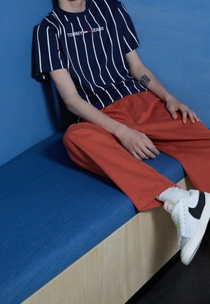 Osoba v námořnicky modrém pruhovaném tričku Tommy Jeans, oranžových kalhotách, bílých ponožkách a bílých teniskách Nike sedící na modré lavičce.