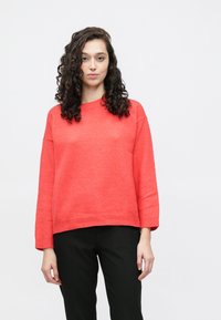 Korallfarget strikkegenser med bred rund hals og senkede skuldre. Har ribbestrikkede mansjetter og kant, kombinert med svarte bukser.