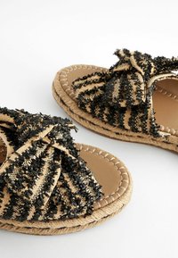 Sandales plates avec semelles en jute tissé et nœuds en tissu rayé noir et naturel sur les brides, présentées sur un fond blanc.