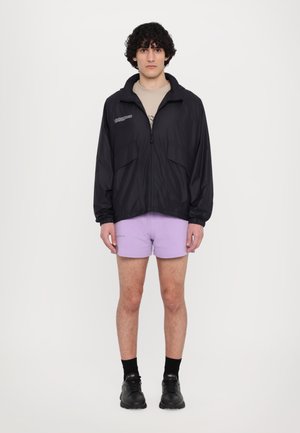 Black lightweight jacket with a zip front, two side pockets, and a high collar. Worn over a beige T-shirt and purple shorts. Black sneakers.