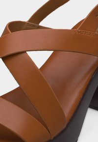 Καφέ δερμάτινο σανδάλι με φαρδιές σταυρωτές λουράκια και μαύρο τακούνι πλατφόρμα, σε κοντινή λεπτομέρεια.