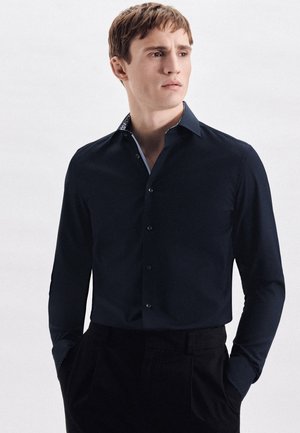 Marineblaues Hemd mit langen Ärmeln und Knopfverschluss, mit gemustertem Innenkragen und glatter Textur. Figurbetonter Schnitt, kombiniert mit dunklen Hosen.