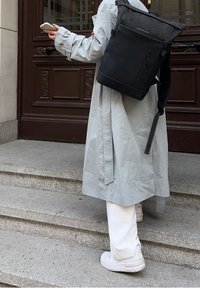 Osoba ubrana w długi jasnoszary płaszcz i białe trampki niesie dużą czarną plecak, trzymając smartfona w pobliżu schodów budynku.