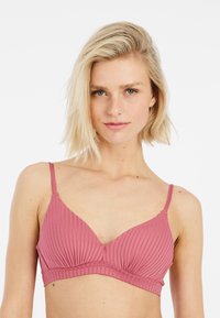 Donna bionda con capelli corti che indossa un reggiseno a coste di colore rosa malva con spalline sottili, rivolta in avanti su uno sfondo bianco.