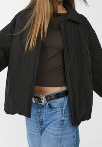 Czarna pikowana kurtka z wysokim kołnierzem, zapinana na zamek, w oversize'owym fasonie, zestawiona z brązową bluzką i niebieskimi dżinsami z wysokim stanem.